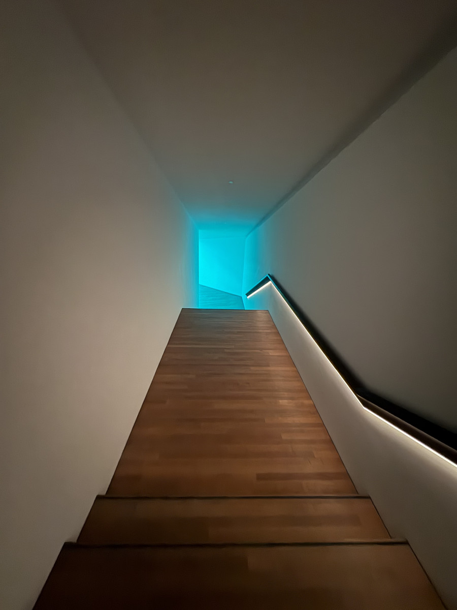 Holztreppe in einem modernen Gebäude, am unteren Ende leuchtet ein blauer Lichtschein.