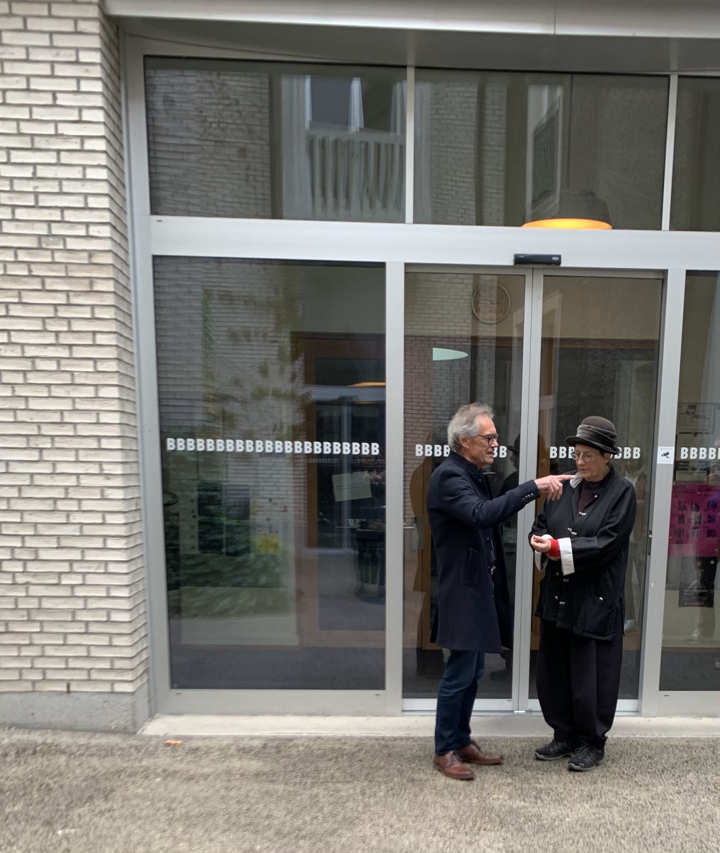 Zwei Personen stehen im Gespräch vor einem Gebäude mit Glasfassade und dem Schriftzug „BBBBBBBB“.