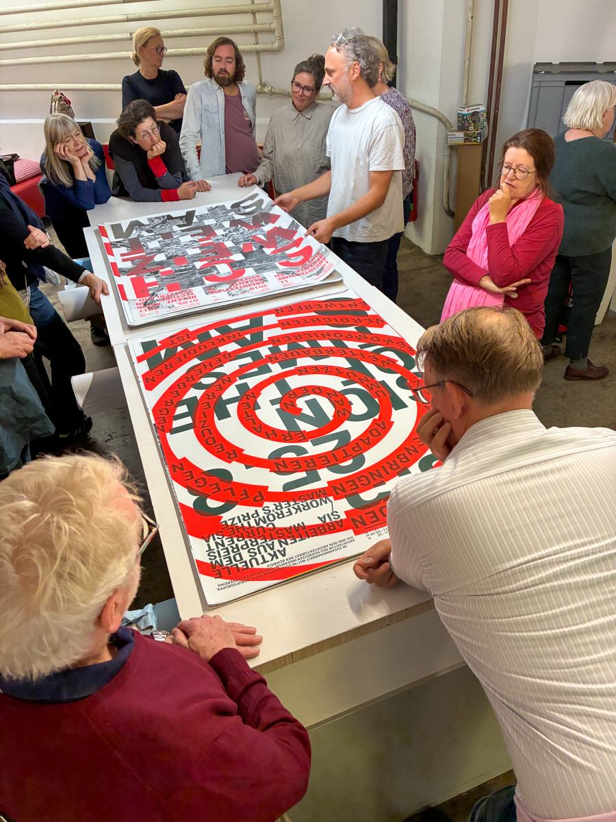 Gruppe von Menschen steht in Jiri Oplateks Atelier um einen Tisch mit großformatigen, grafisch gestalteten Plakaten in Rot und Schwarz.