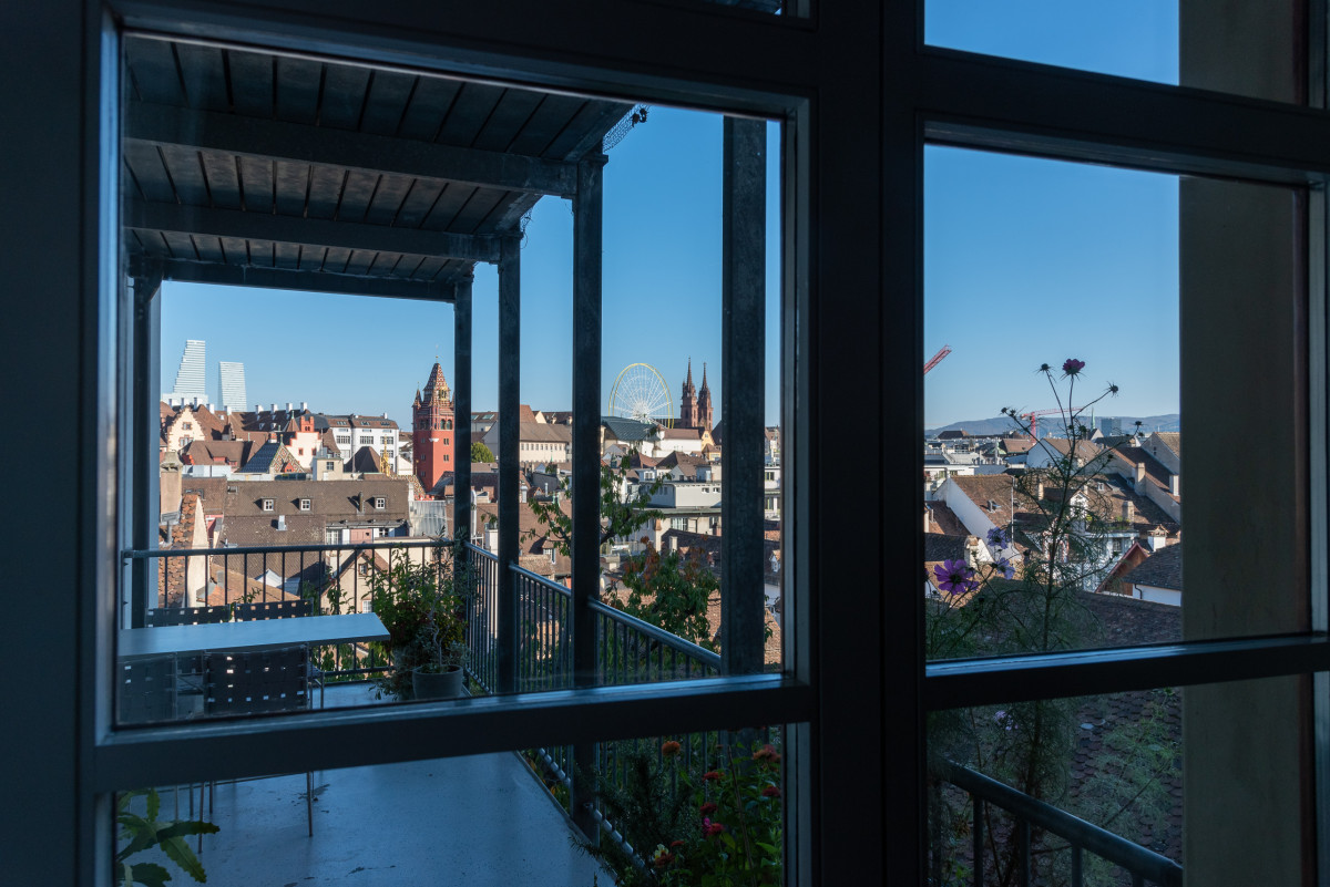 Blick durch eine Glasfront auf Basel: Terrasse mit Blumen, dahinter Altstadtdächer, Münster und Riesenrad.