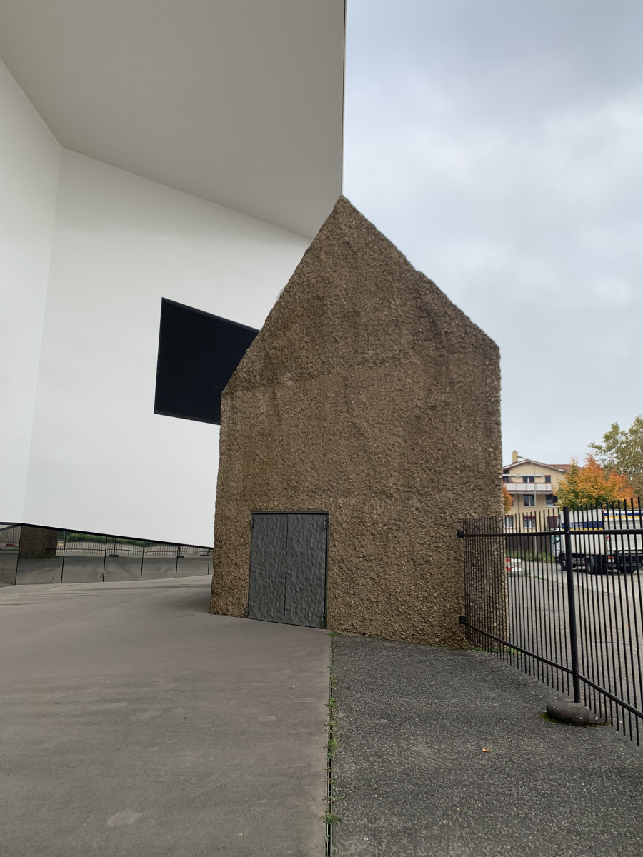 Kleines, grob verputztes Haus steht neben einem großen weißen Museumsbau.