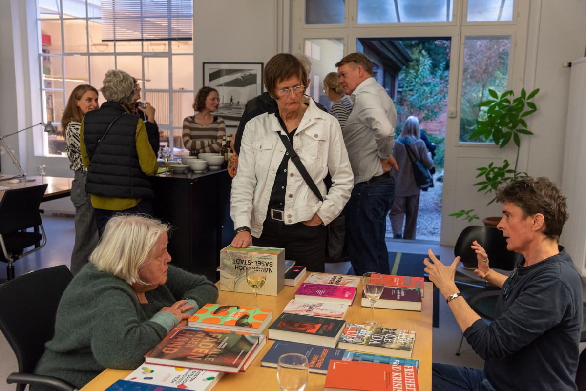 Menschen unterhalten sich in einem Raum, auf dem Tisch liegen zahlreiche Bücher und Weingläser.
