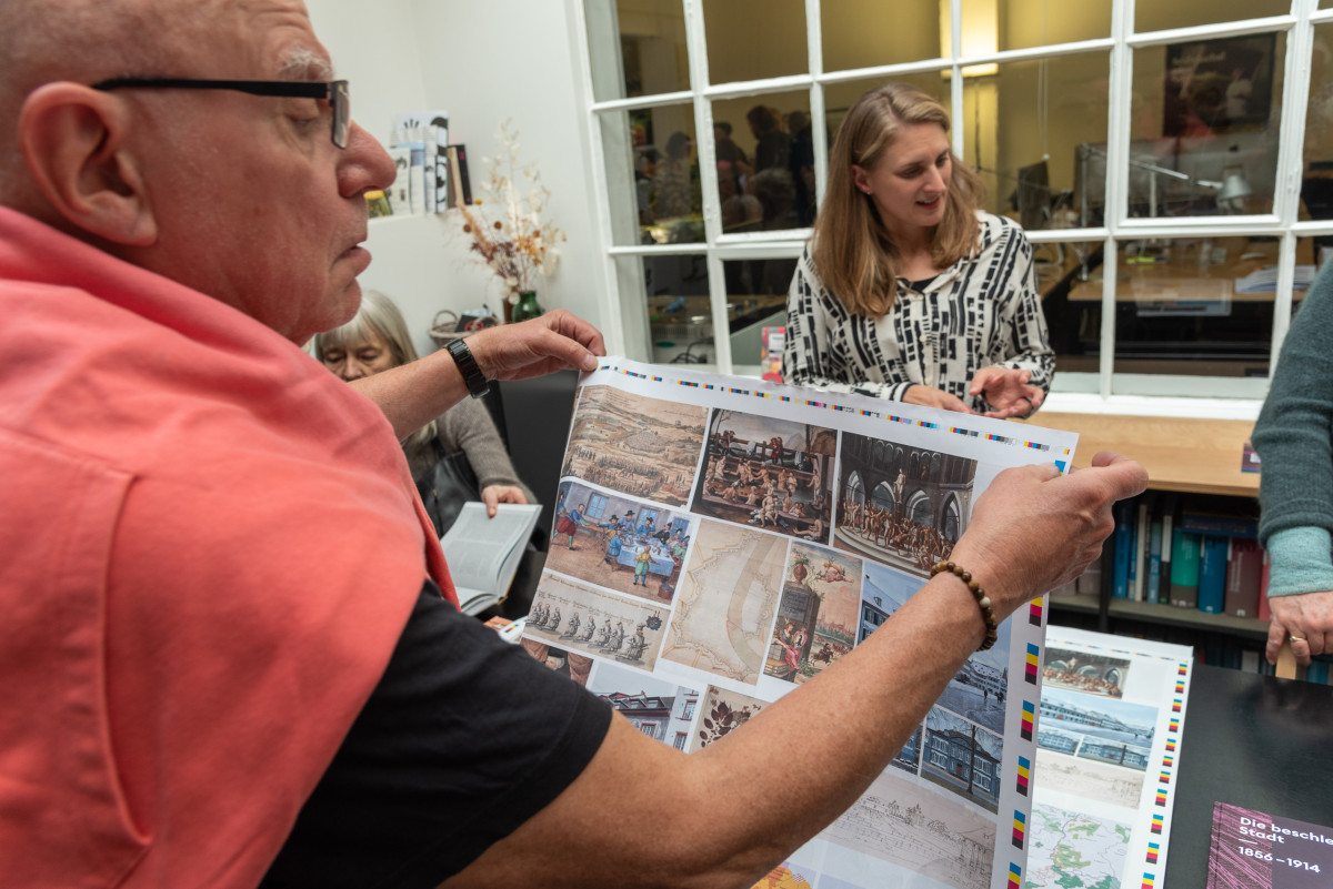 Person hält einen Druckbogen, andere betrachten Bücher und Druckproben.