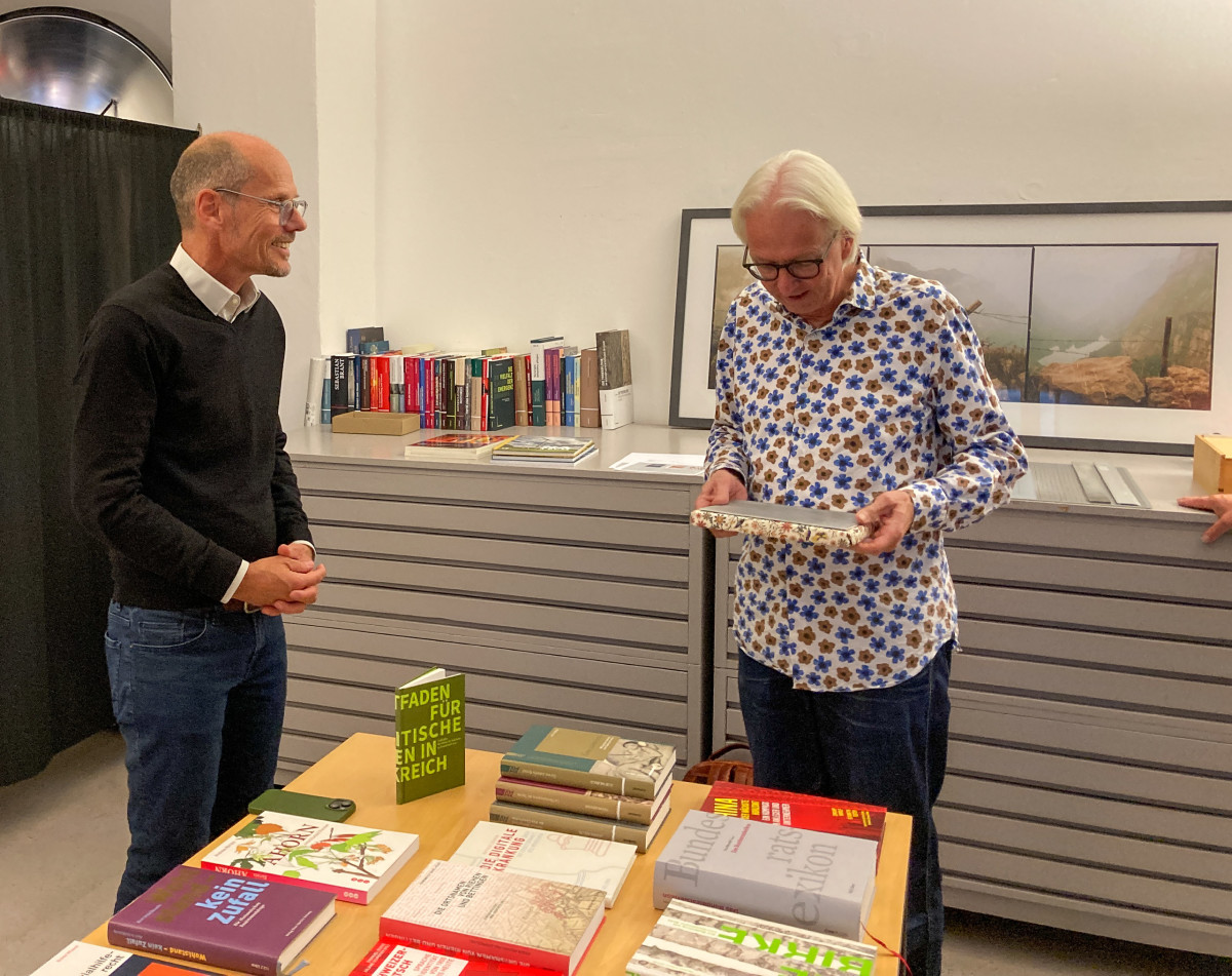 Zwei Personen stehen an einem Tisch voller Bücher, Christoph Draxler betrachtet ein Buch.