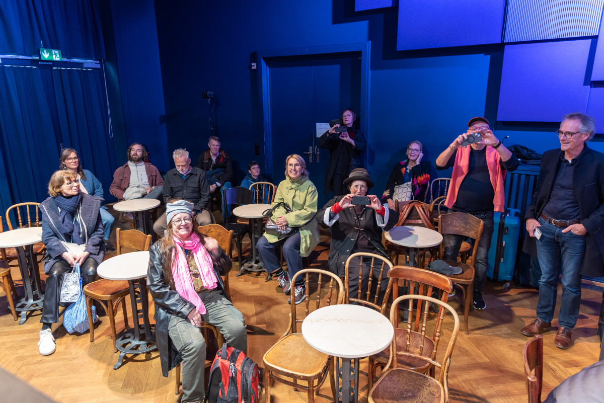 Gruppe von Menschen sitzt in einem kleinen blauen Saal mit Caféhausmöbeln und lacht.