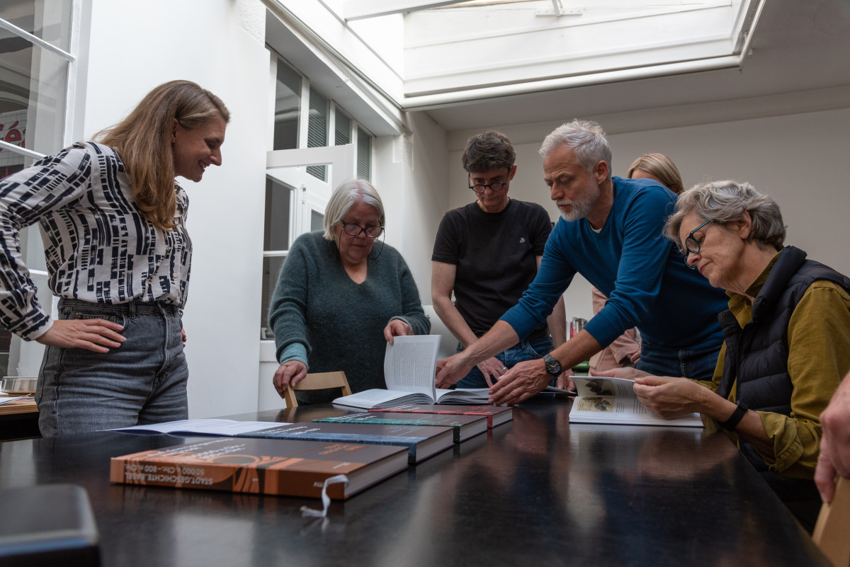 Gruppe von Personen blättert gemeinsam in Büchern und diskutiert an einem Tisch in einem hellen Raum.