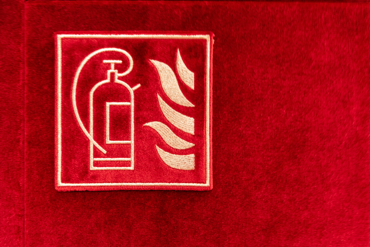 Piktogramm eines Feuerlöschers mit Flammen, in goldener Stickerei auf rotem Samtuntergrund.
