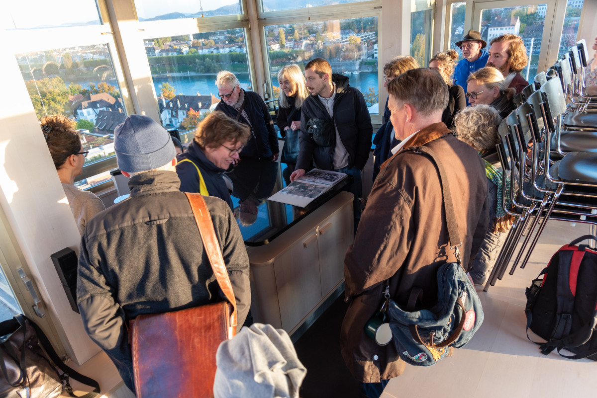 Gruppe steht im lichtdurchfluteten Raum mit Blick über den Rhein, jemand zeigt ein offenes Buch.