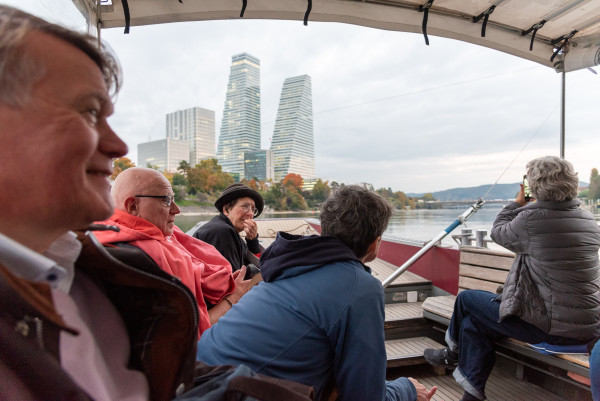Gruppe von Personen sitzt auf einem Boot auf dem Rhein, im Hintergrund die Roche-Türme von Basel.