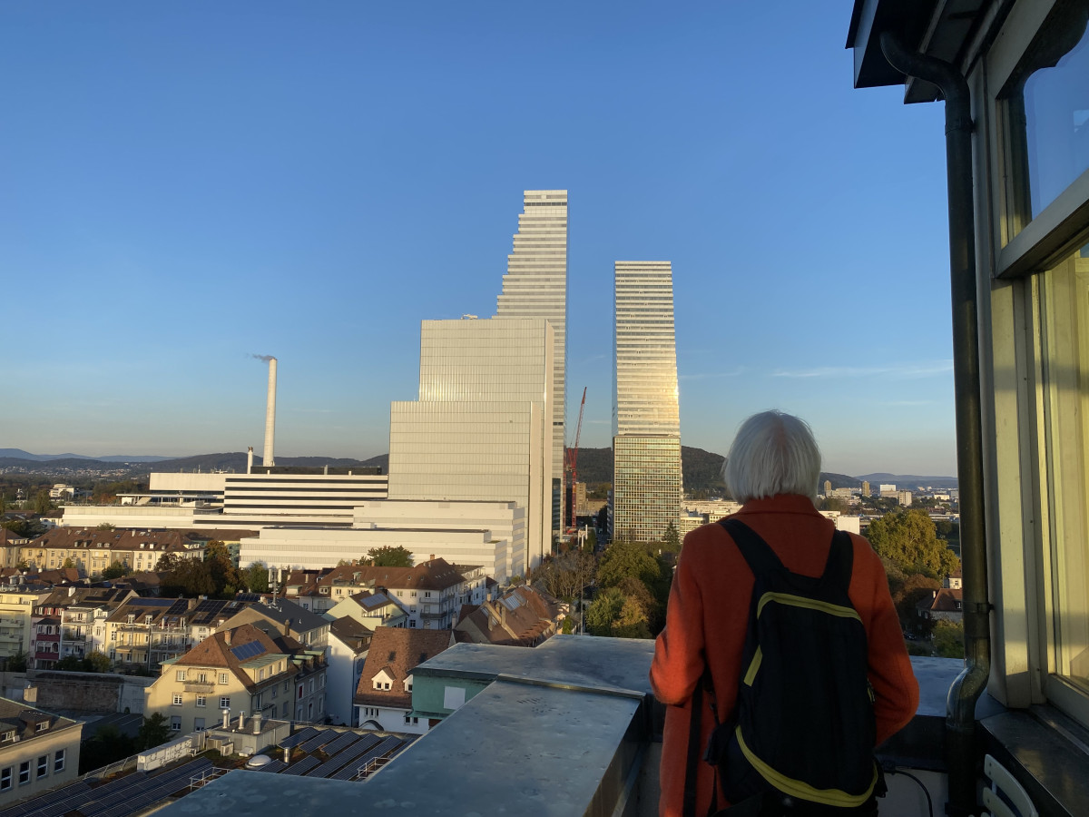 Christoph Draxler mit orangefarbener Jacke blickt vom Dach aus auf die modernen Roche-Türme von Basel.