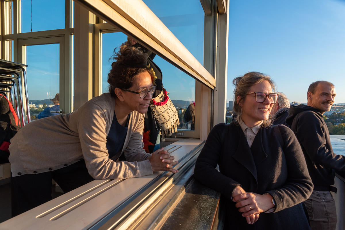 Zwei Teilnehmerinnen lehnen an einer Fensterfront mit Blick auf die Stadt, warmes Abendlicht fällt ins Innere.