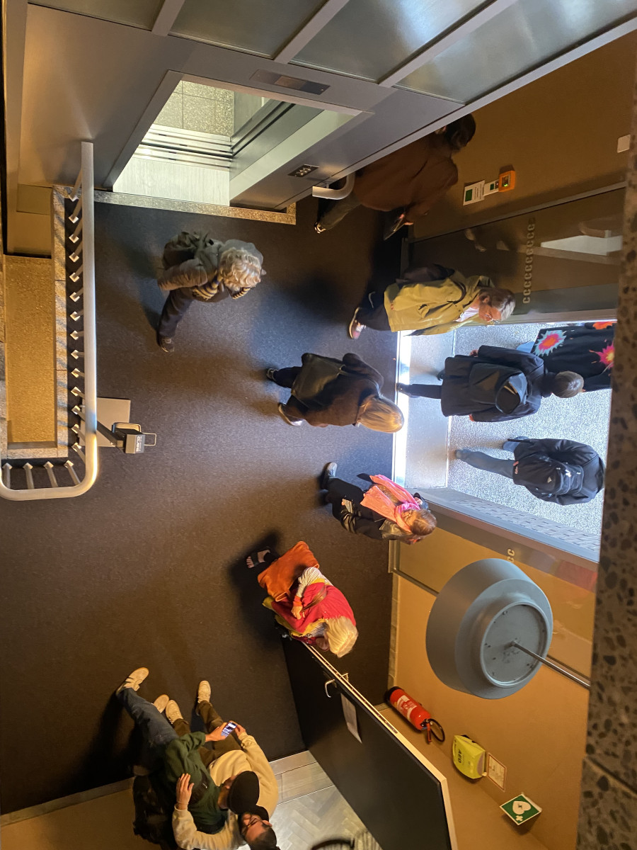 Blick von oben auf Menschen, die durch einen Eingangsbereich mit Fahrstuhl gehen.