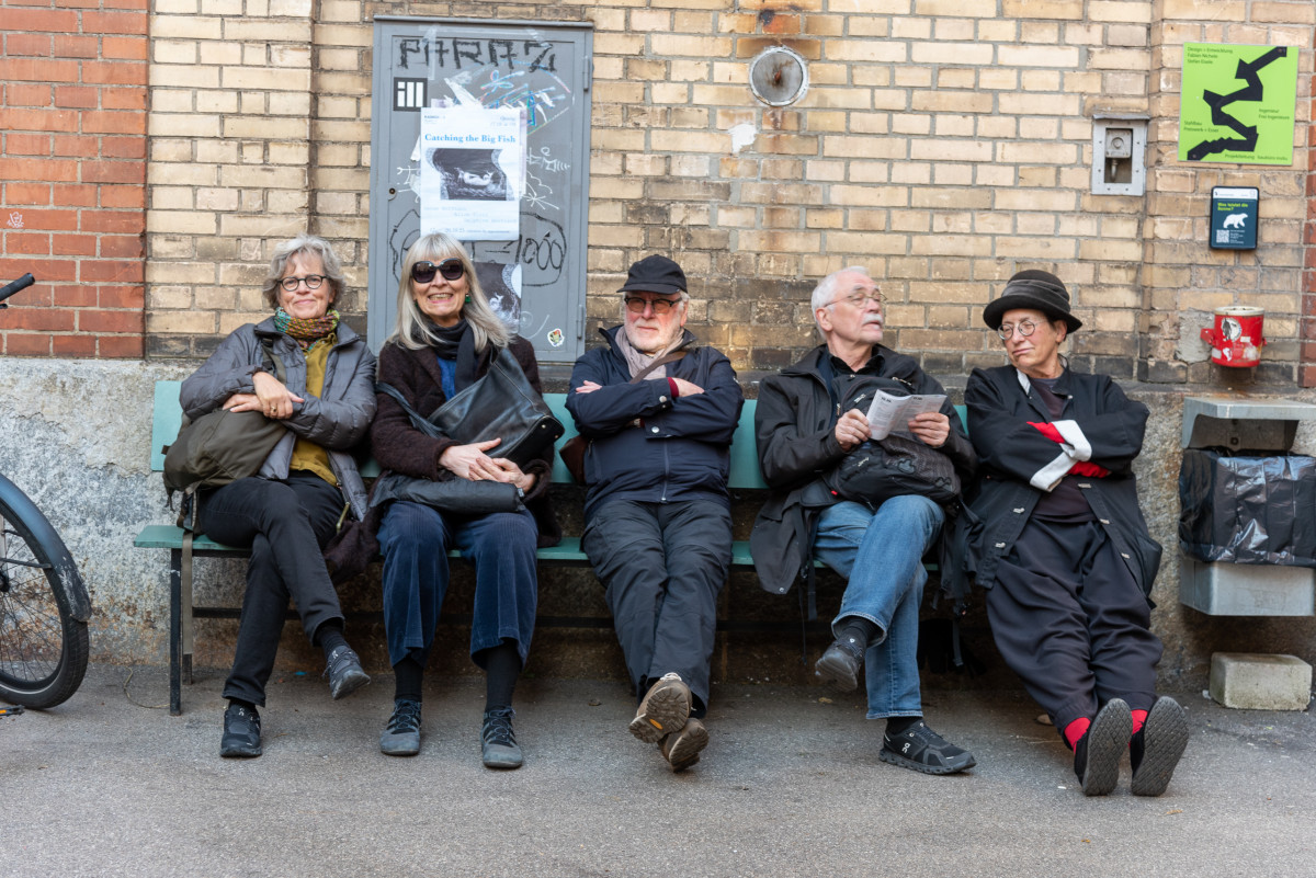 Fünf Personen sitzen auf einer Bank vor einer Backsteinwand, lachen und unterhalten sich.