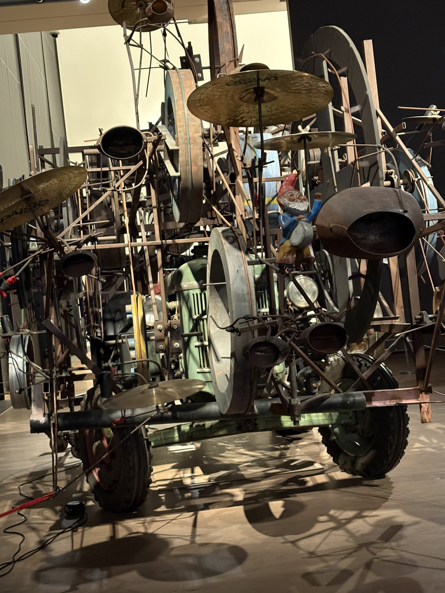 Komplexe mechanische Skulptur aus Metallteilen, Trommeln und Becken in einem Ausstellungsraum.