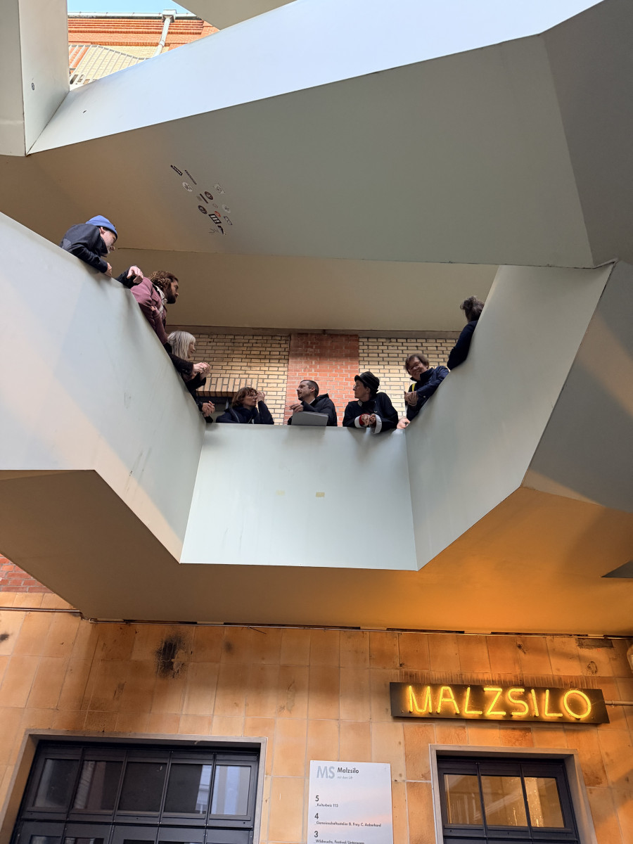 Gruppe blickt von einem Balkon unter markanten Betonträgern nach unten. Darunter: Ein Schild „Malzsilo“ leuchtet in Gelb.