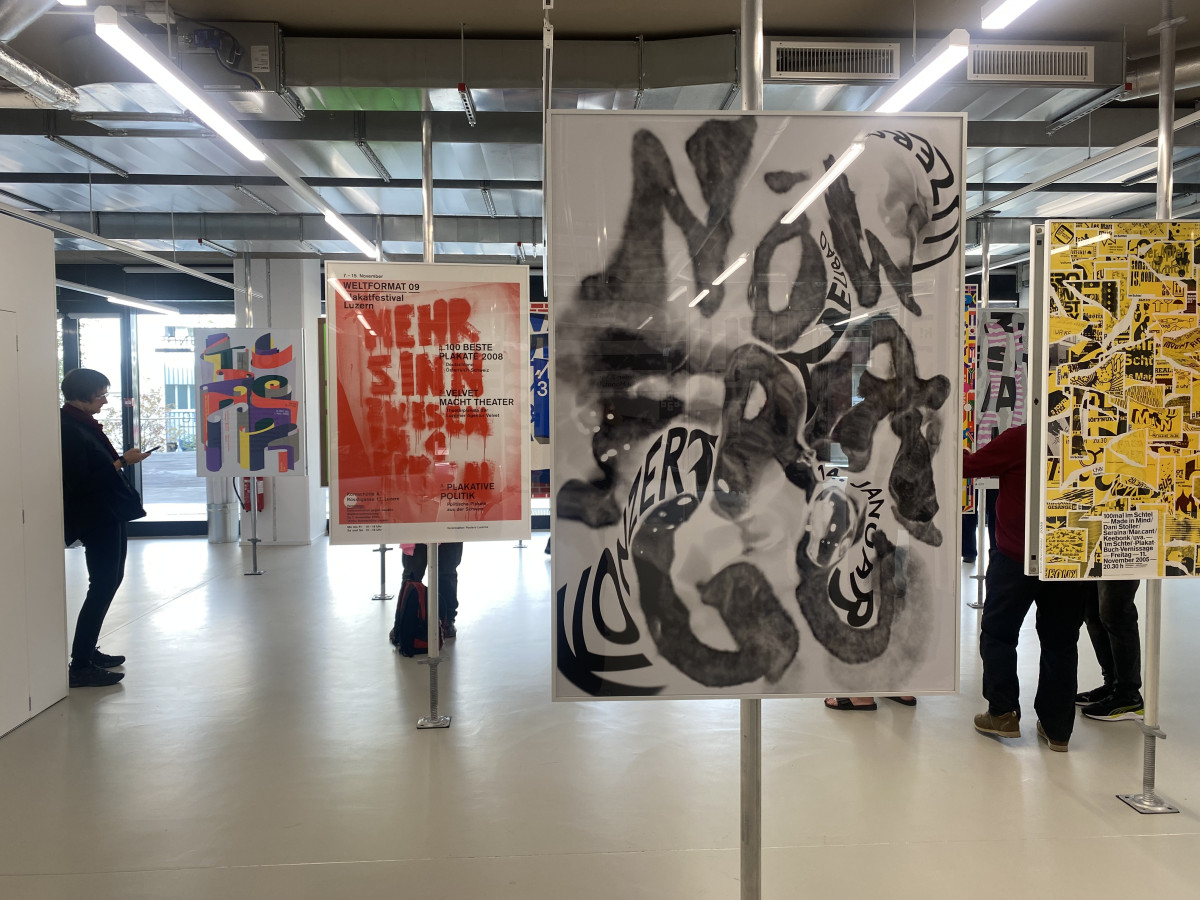 Ausstellung mit Plakaten in verschiedenen Stilen und Farben, Besucherinnen und Besucher bewegen sich durch den Raum.