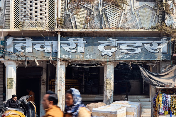 Die Fassade eines weißen Art-déco-Gebäudes mit großen Kisten auf den Stufen und einem Paar, das auf einem Zweirad davor vorbeifährt. Das Gebäude hat ein Schild in Graublau, auf dem in Devanagari »Tiwari Traders« steht.