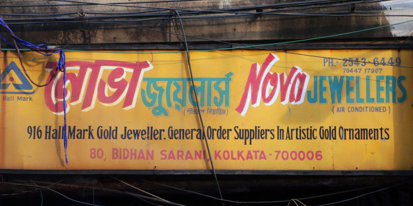 Ein orangefarbenes Schild an einem baufälligen braunen Gebäude. Auf dem Schild steht „Nova Jewellers“ in Bengali und lateinischer Schrift, außerdem sind die Dienstleistungen und die Adresse des Geschäfts angegeben.