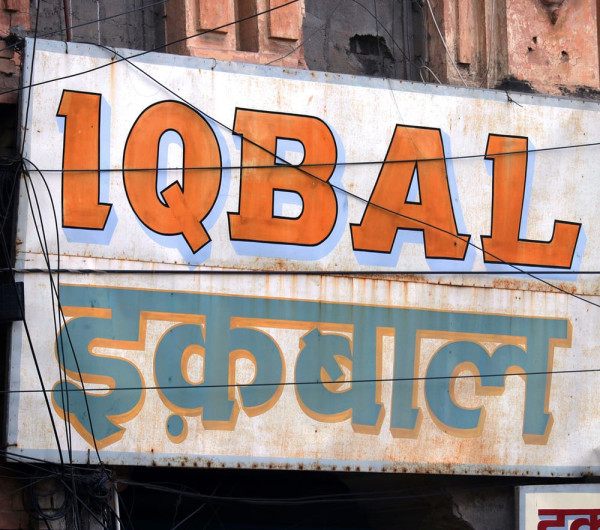 Ein weißes Schild mit einem schmalen blauen Rand, auf dem »Iqbal« in lateinischer Schrift und Devanagari steht. Die Devanagari-Schrift übernimmt die Serifen des Textes in lateinischer Schrift.