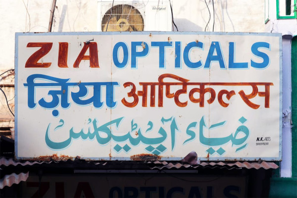 Ein weißes Schild mit einem schmalen blauen Rand, auf dem in aufeinanderfolgenden Zeilen »Zia Opticals« in lateinischer Schrift, Devanagari und Nastaliq steht.