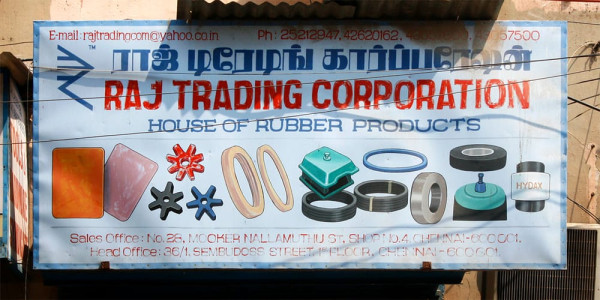 Mehrsprachiges Schild für Raj Trading Corporation mit tamilischem Text in Blau und lateinischem Text in Rot sowie Abbildungen von Gummiprodukten, die das Geschäft verkauft.