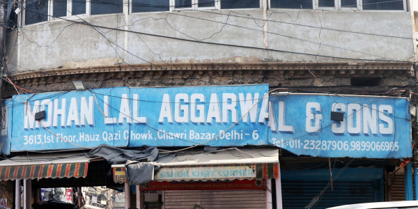 Die Fassade eines grauen Gebäudes mit großen Fenstern und einem breiten blauen Schild mit der Aufschrift »Mohanlal Agarwal & Sons« sowie der Adresse des Gebäudes und den Telefonnummern des Unternehmens.