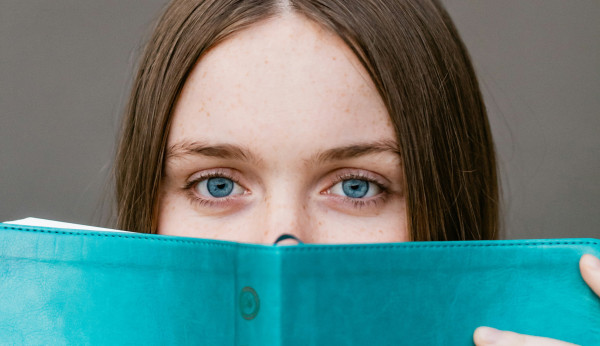 eine junge Erwachsene schaut über den Rand eines Buches in die Kamera
