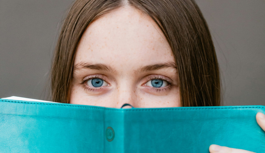 eine junge Erwachsene schaut über den Rand eines Buches in die Kamera