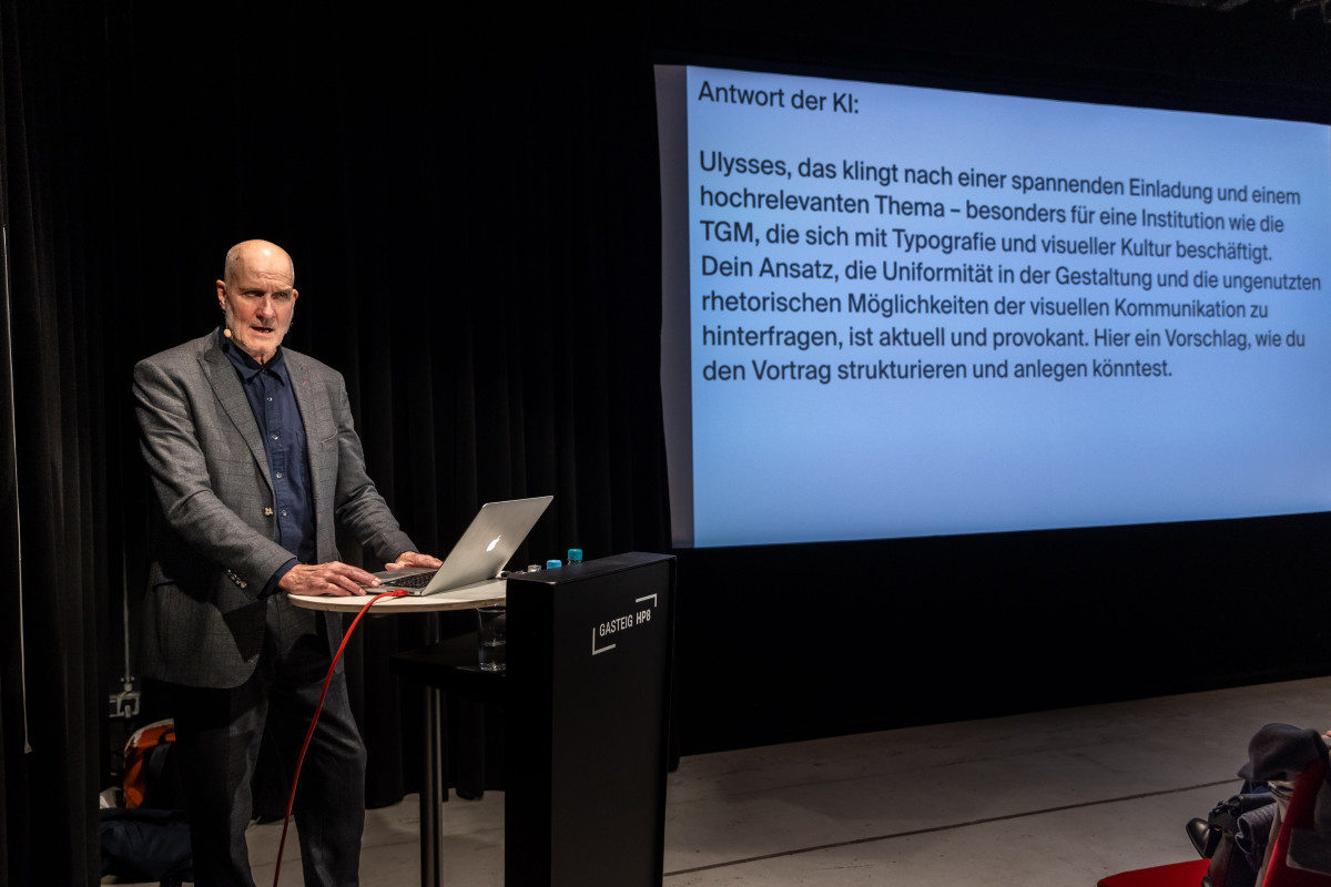 Ulysses Voelker zeigt eine Präsentationsfolie mit dem Text: „Antwort der KI: Ulysses, das klingt nach einer spannenden Einladung und einem hochrelevanten Thema - besonders für eine Institution wie die TGM, die sich mit Typografie und visueller Kultur beschäftigt. Dein Ansatz, die Uniformität in der Gestaltung und die ungenutzten rhetorischen Möglichkeiten der visuellen Kommunikation zu hinterfragen, ist aktuell und provokant. Hier ein Vorschlag, wie du den Vortrag strukturieren und anlegen könntest.“