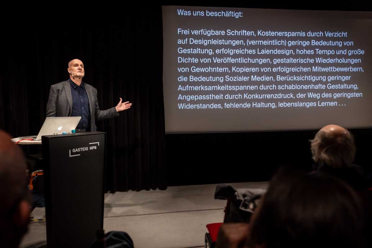 Prof. Jean Ulysses Voelker spricht an einem Podium vor Laptop und Präsentationsfolie („Was uns beschäftigt:“), gestikuliert und analysiert kritisch Herausforderungen im Gestaltungsberuf wie Vereinheitlichung, Tempo und fehlende Haltung, mit zugleich ermutigendem Ausblick.