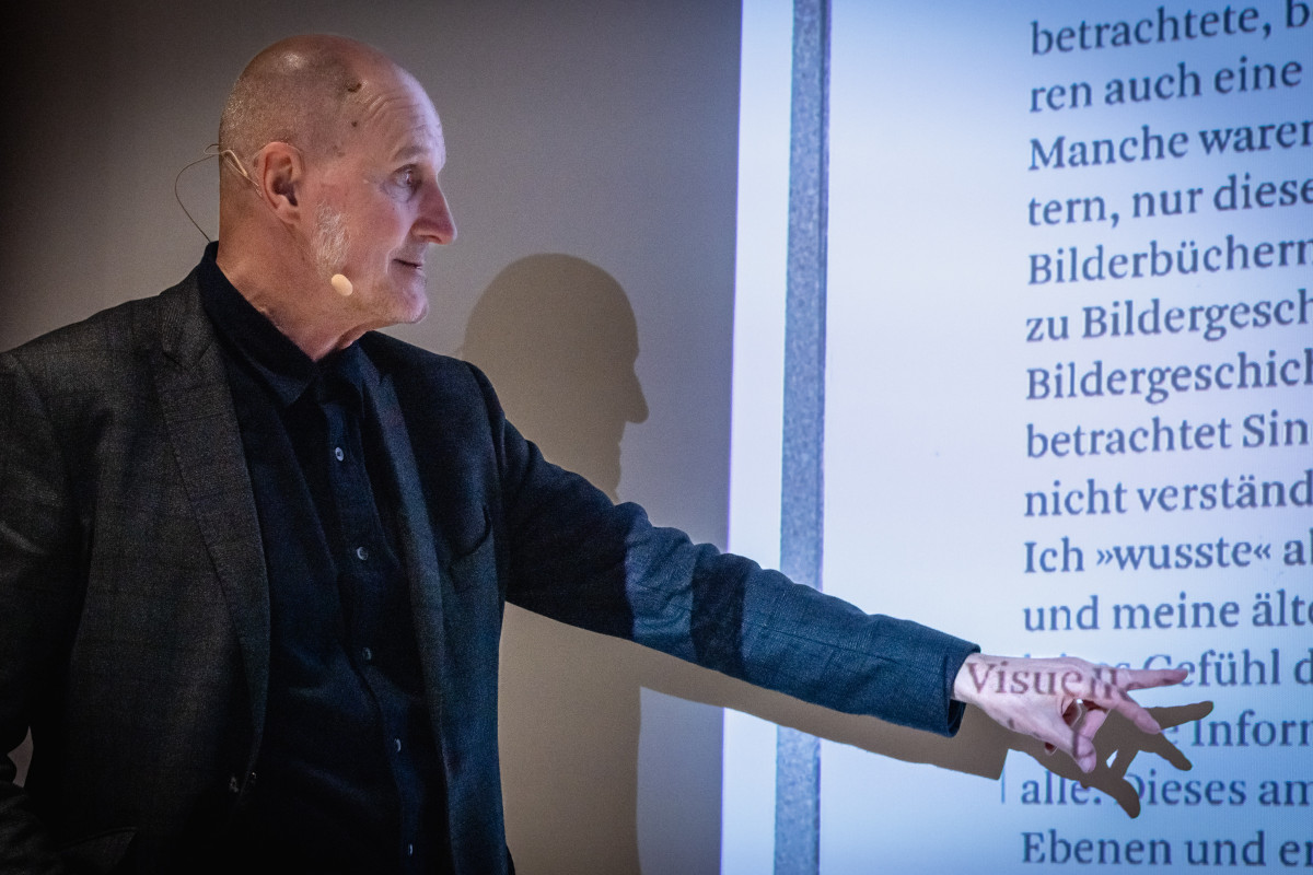 Prof. Jean Ulysses Voelker als Eröffnungsredner der tgm-Reihe „Zeichen setzen – Gestaltung braucht Haltung“, zeigt auf eine Projektionsfolie zu visueller Rhetorik und reflektiert grundlegende Prinzipien der visuellen Kommunikation.
