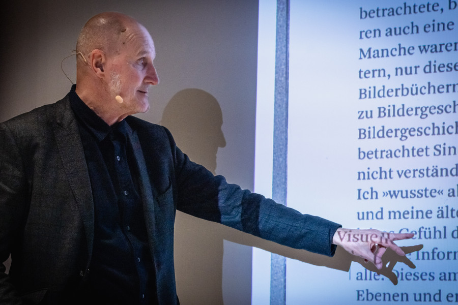 Prof. Jean Ulysses Voelker als Eröffnungsredner der tgm-Reihe „Zeichen setzen – Gestaltung braucht Haltung“, zeigt auf eine Projektionsfolie zu visueller Rhetorik und reflektiert grundlegende Prinzipien der visuellen Kommunikation.